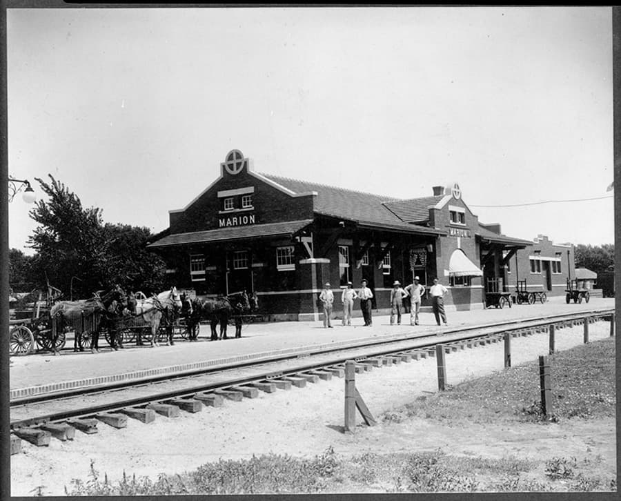Historic black and white image of the Marion train station with horse-drawn wagons and people gathered outside.