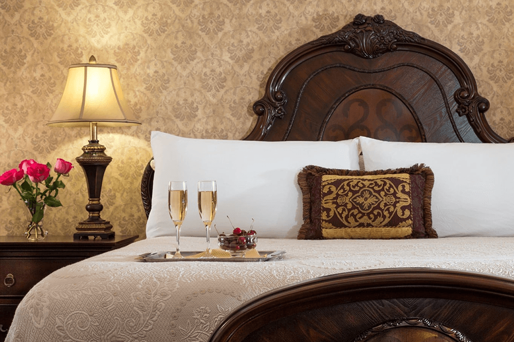 A beautifully arranged bedroom featuring a bed with ornate headboard, a lamp, and glasses of champagne on a tray.