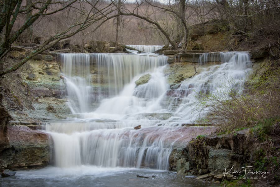 A tranquil waterfall cascades over rocky ledges surrounded by bare trees.