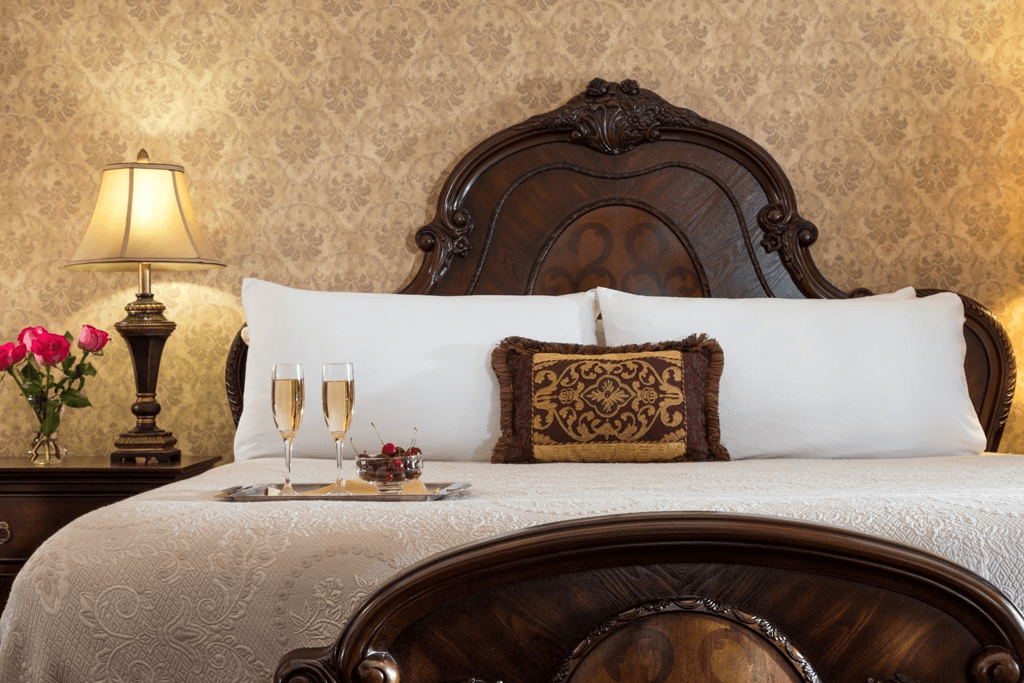 A luxurious bedroom featuring an ornate wooden headboard, soft white bedding, and a tray with champagne and fruit.