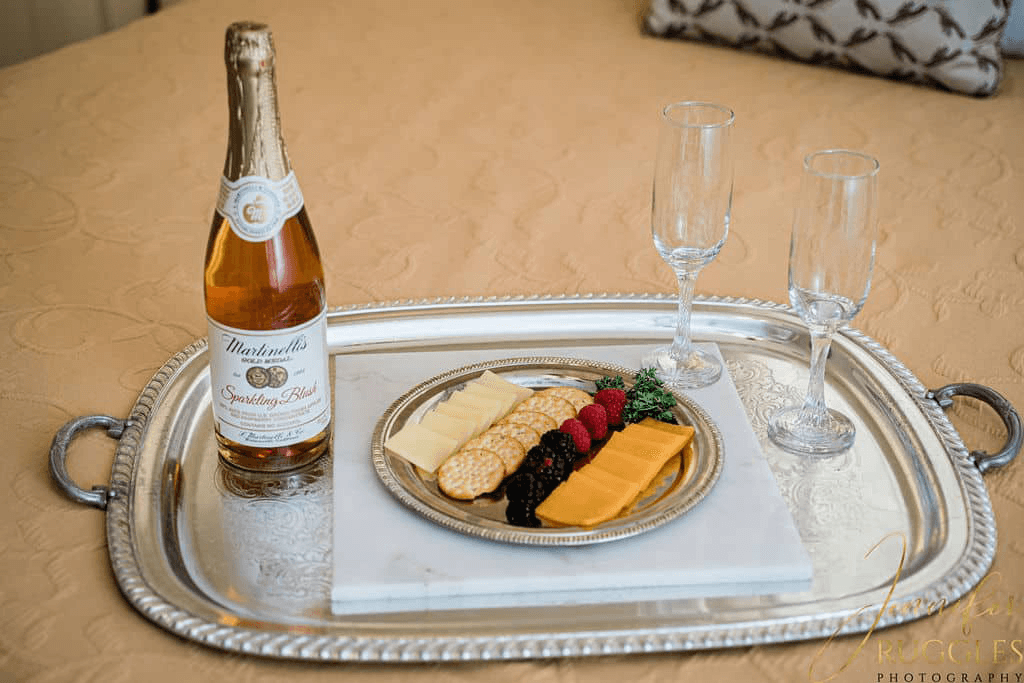 A silver tray featuring a bottle of sparkling cider, two flutes, and a platter of assorted cheeses and berries.