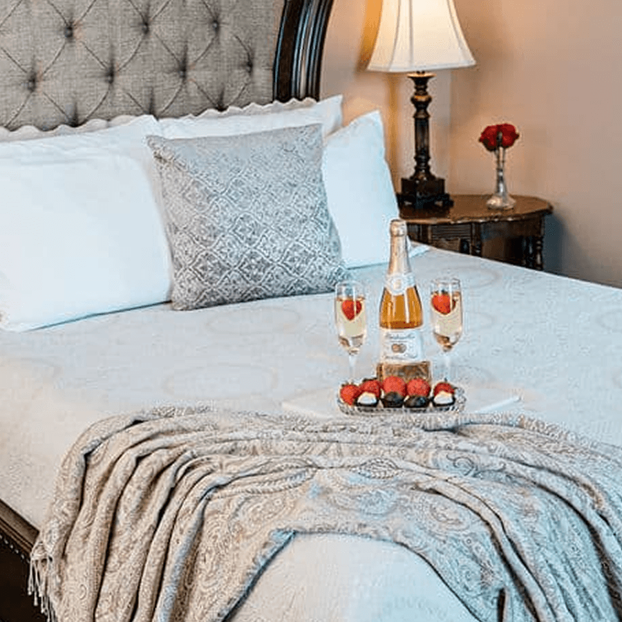 A neatly arranged bed with pillows, a cozy blanket, and a tray of champagne, strawberries, and chocolate-covered treats.