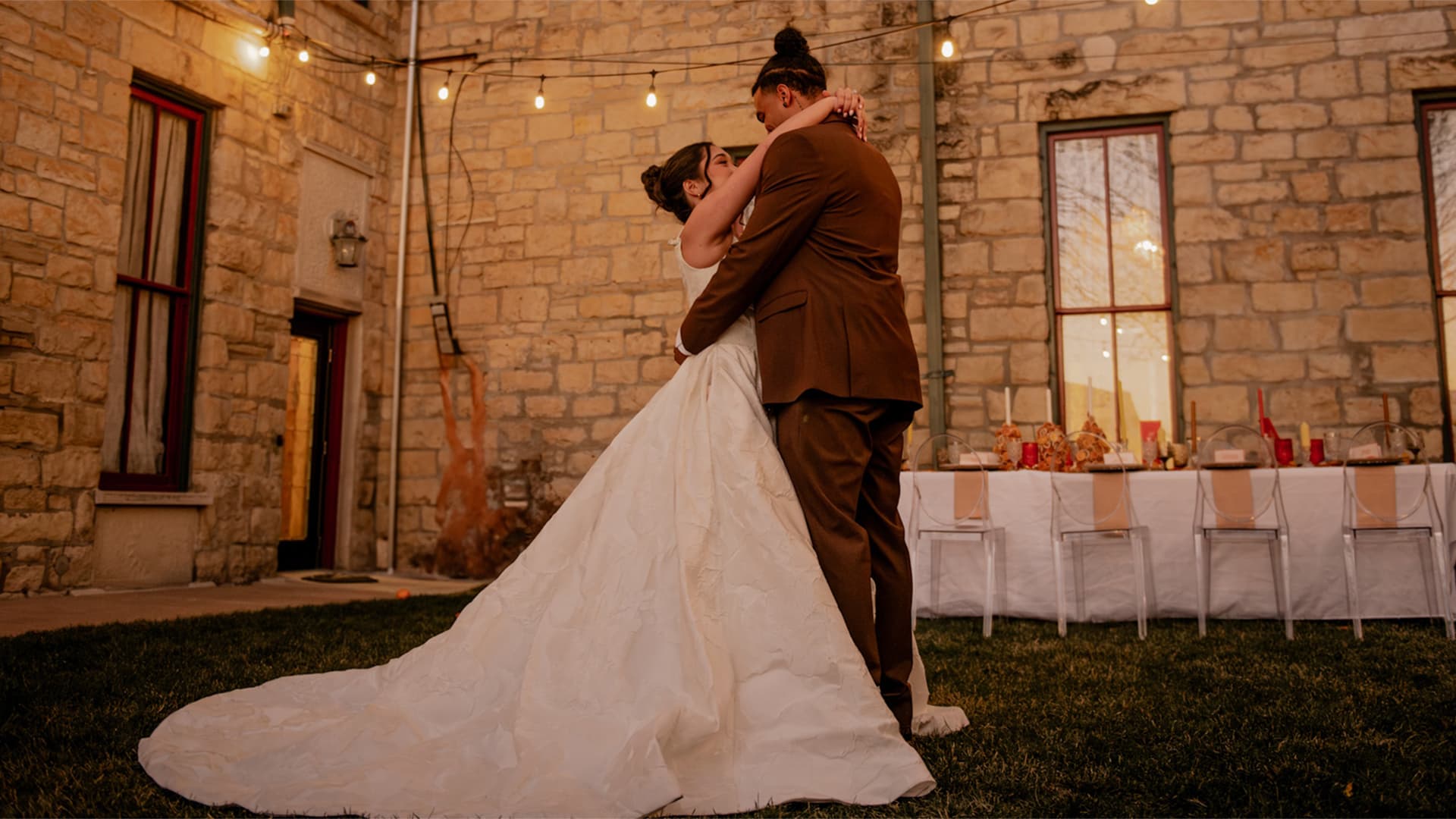A couple shares a romantic embrace outdoors, surrounded by warm lights and elegant decor.