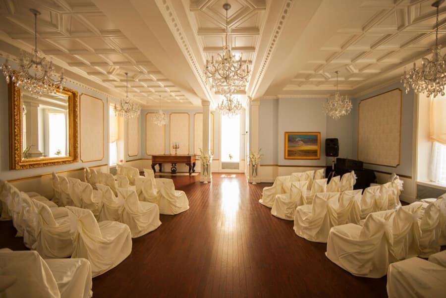 A beautifully decorated event room with white-covered chairs, elegant chandeliers, and a large mirror.