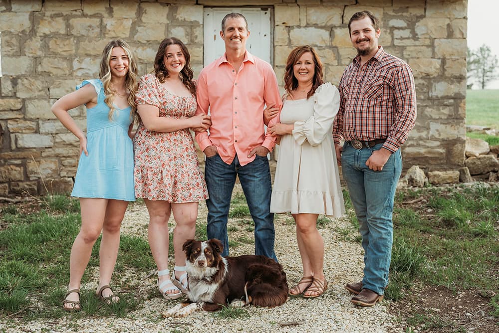 A family poses together outdoors with a dog in front of a rustic stone building.
