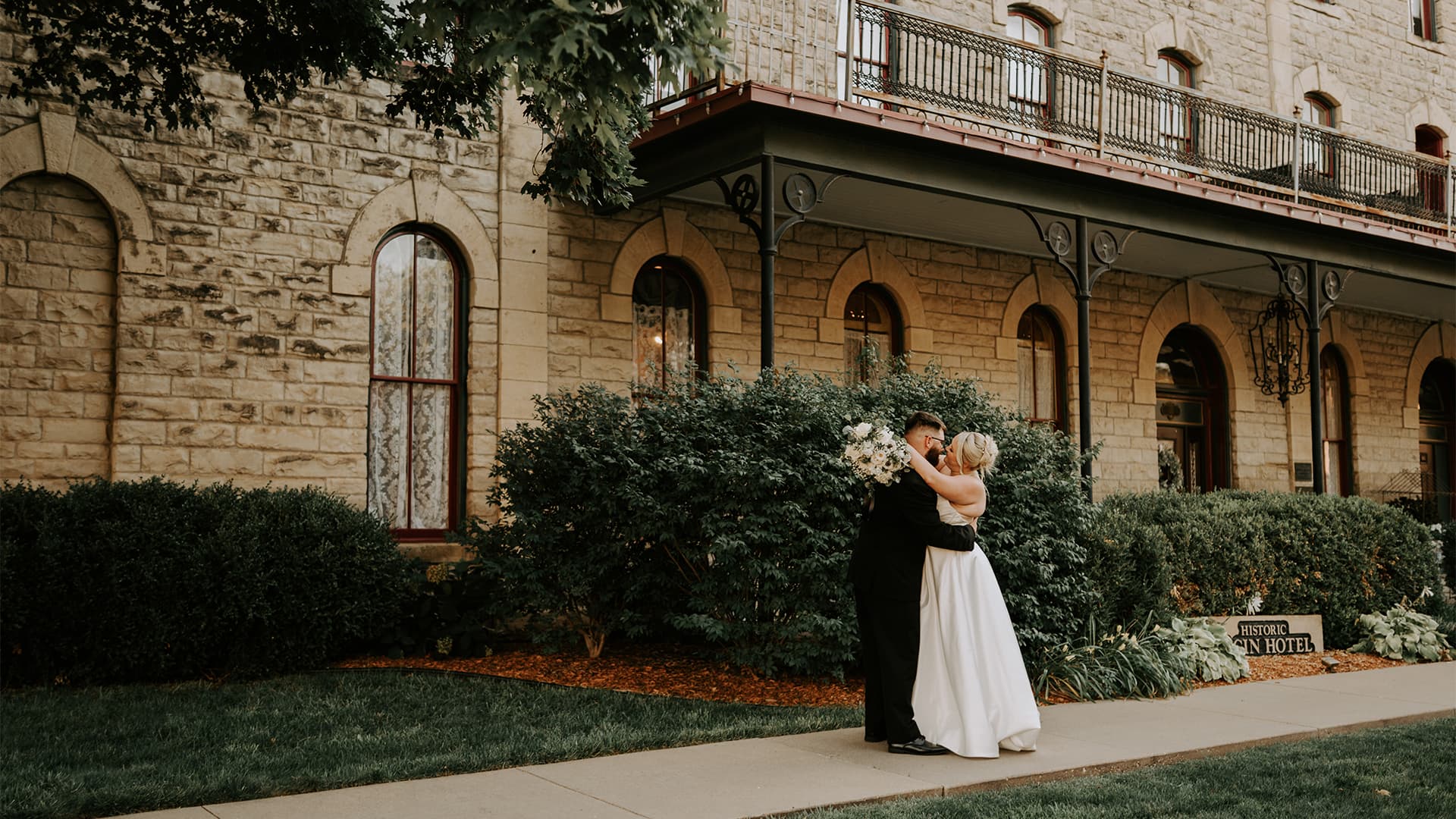 A couple embraces in front of a historic stone building, surrounded by greenery.