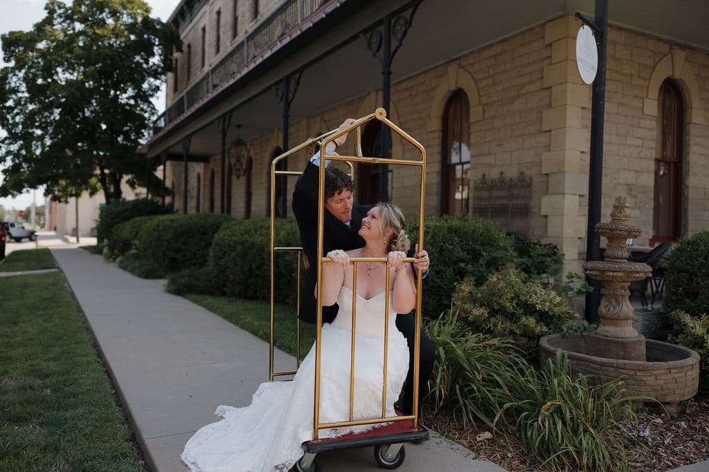 A bride playfully sits in a luggage cart outside a building while a friend assists her.