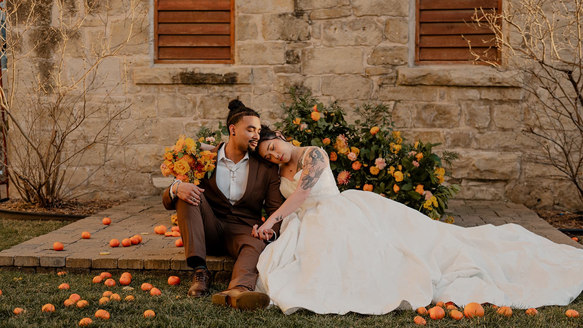 A joyful couple sits together on a stone platform, surrounded by orange blossoms and scattered citrus fruits.