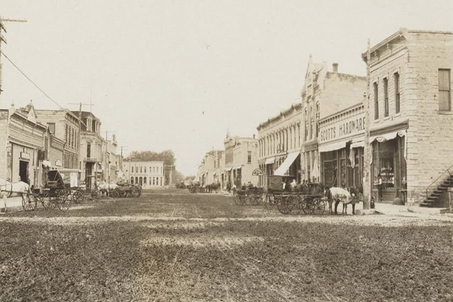 A historical sepia-toned photo of a bustling street lined with horse-drawn carriages and storefronts.
