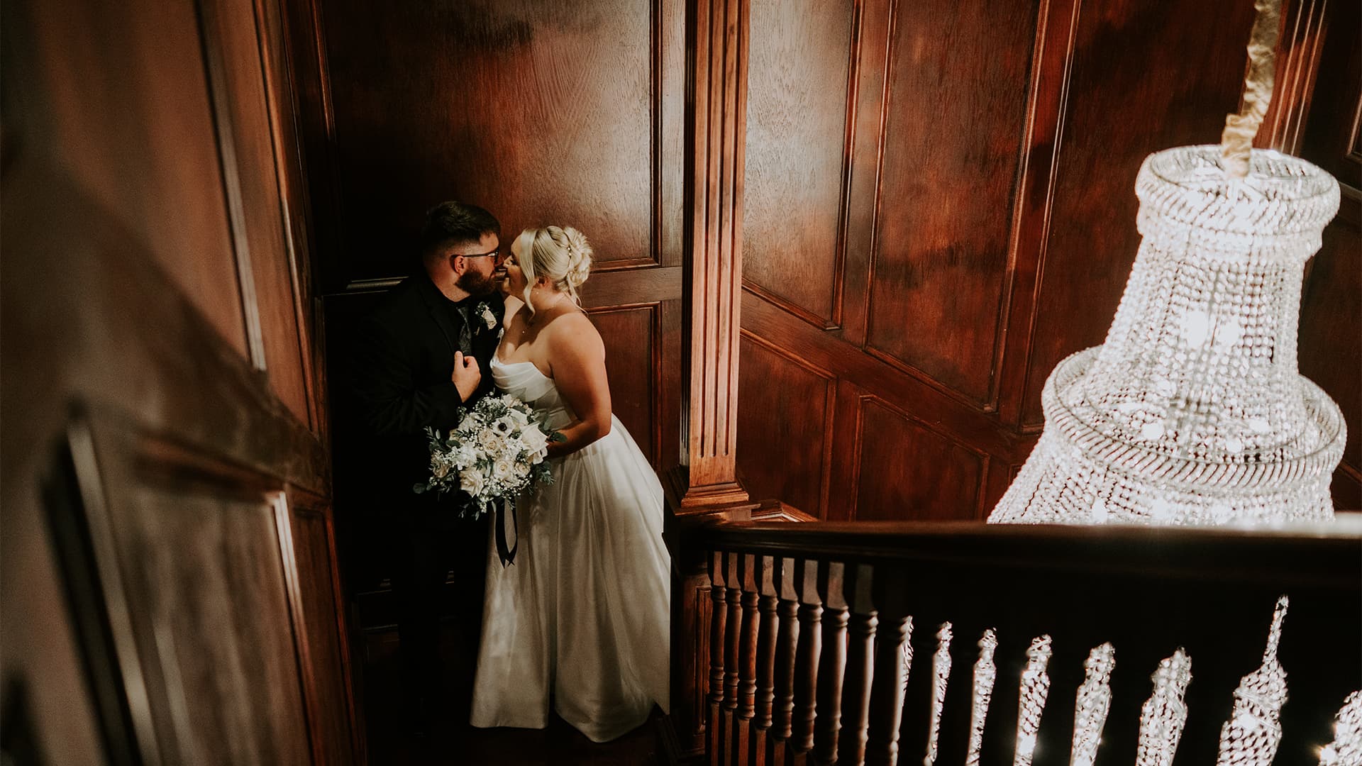 A bride and groom share a romantic moment on a staircase adorned with a chandelier.