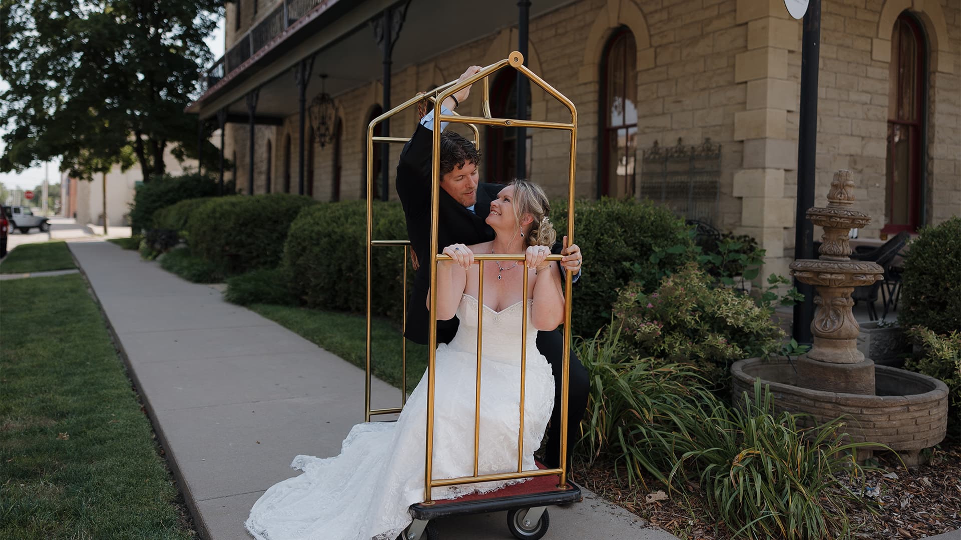 A bride and groom playfully pose inside a luggage cart outside a historic building.