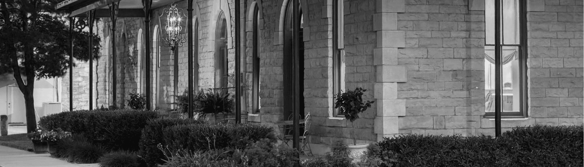 A black-and-white image of a stone building with large windows and lush greenery in front.
