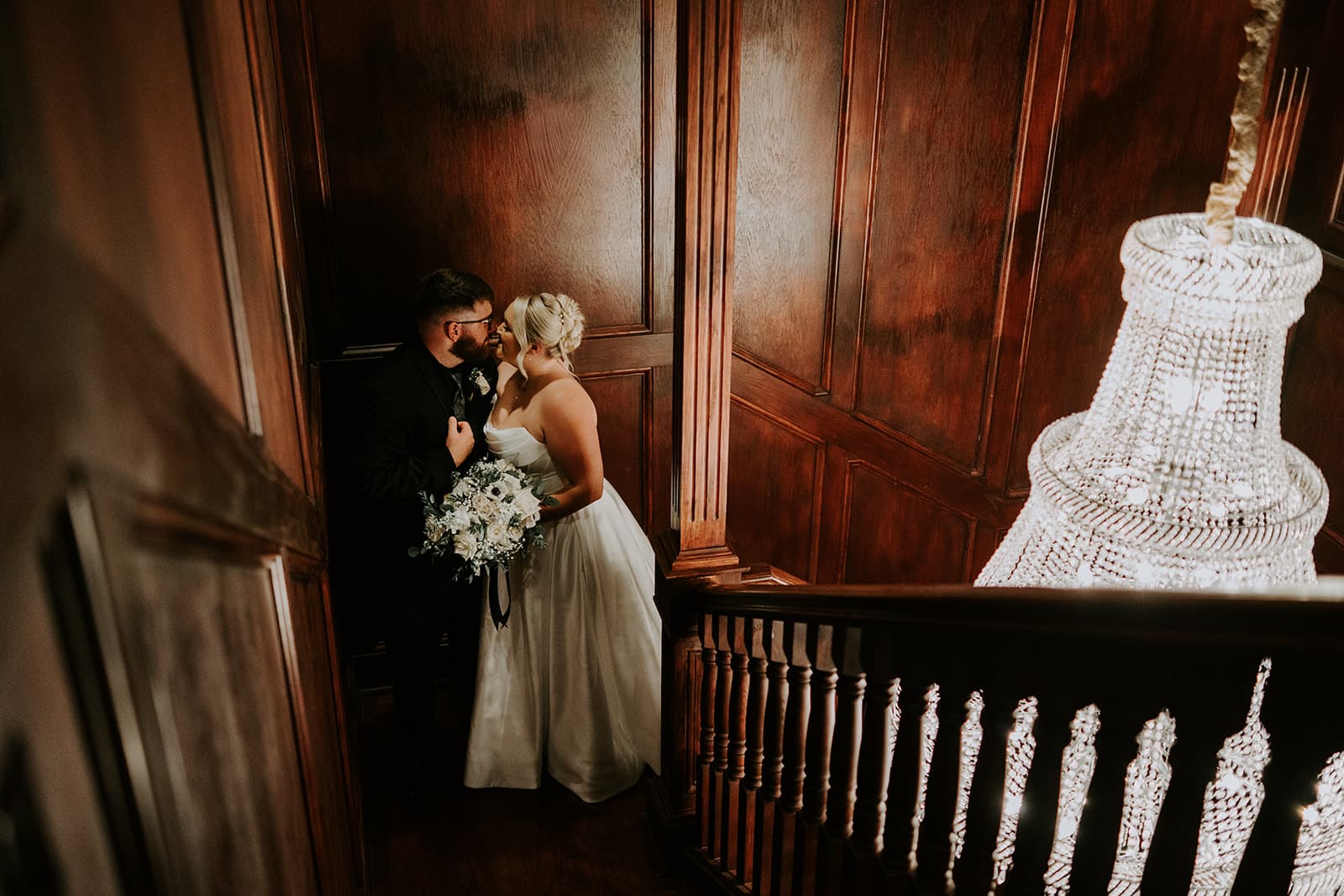 A couple shares a romantic moment on a staircase adorned with a chandelier.