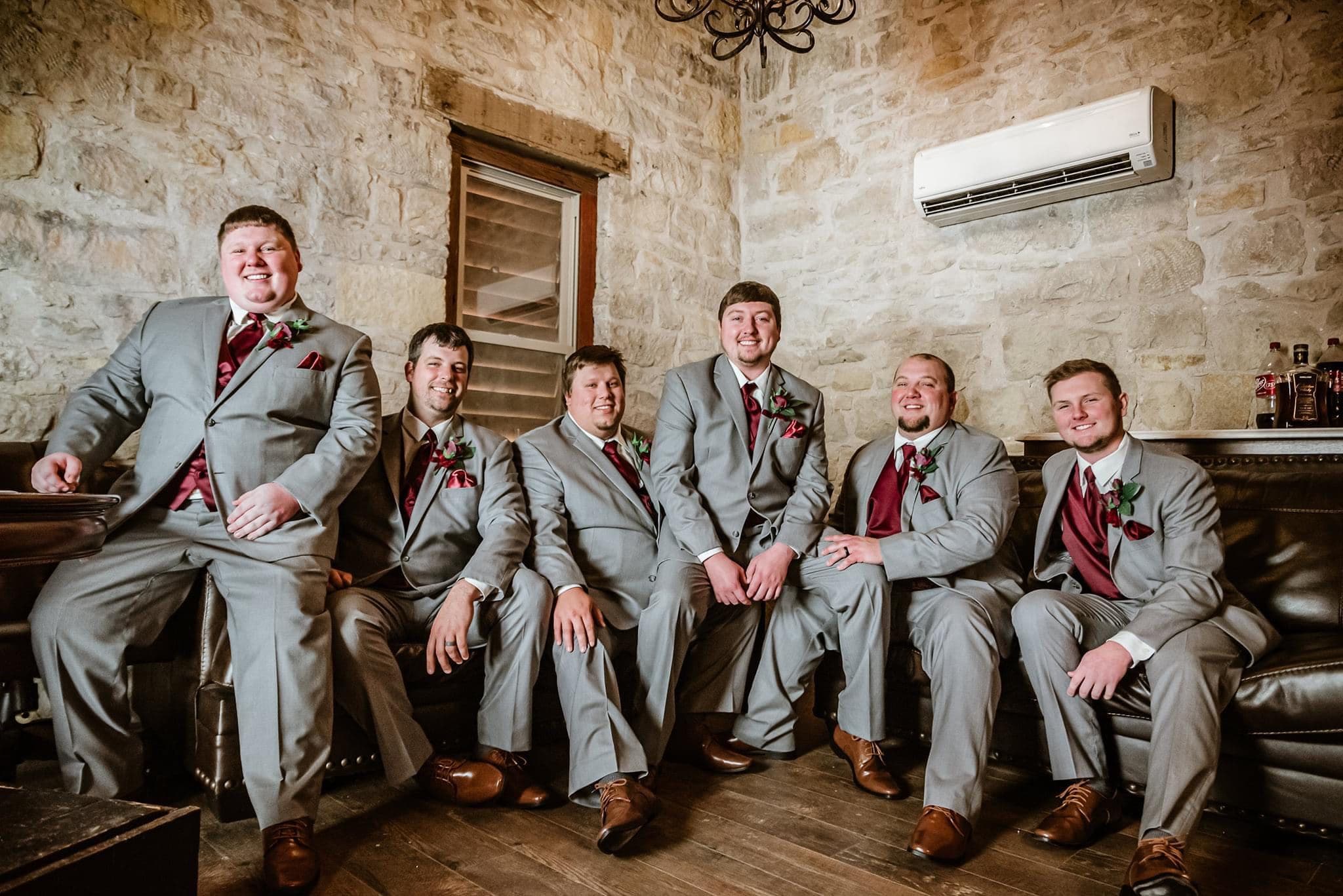 A group of six men in gray suits with maroon accents smiles together in a cozy, stone-walled setting.