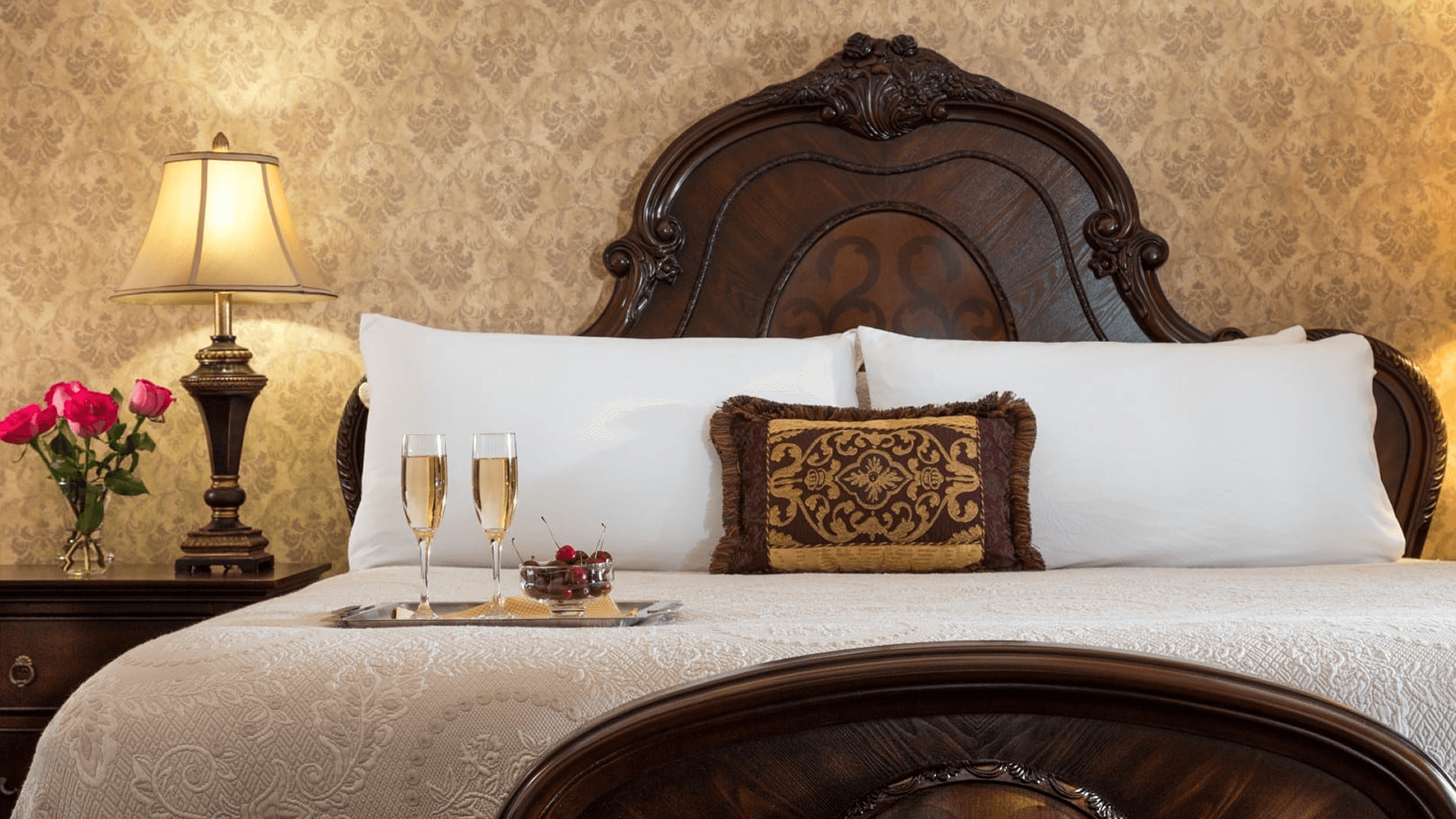 Elegant guest suite at Historic Elgin Hotel in Marion, Kansas, featuring a vintage carved wood bed with plush bedding, fresh flowers, and a romantic champagne tray.