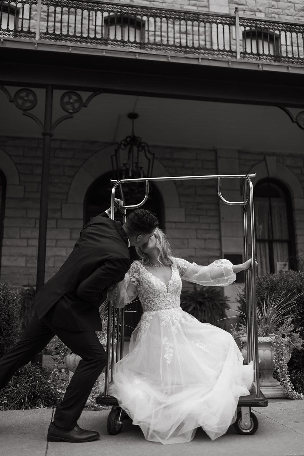 A couple kisses playfully while sitting in a luggage cart outside a historic building.