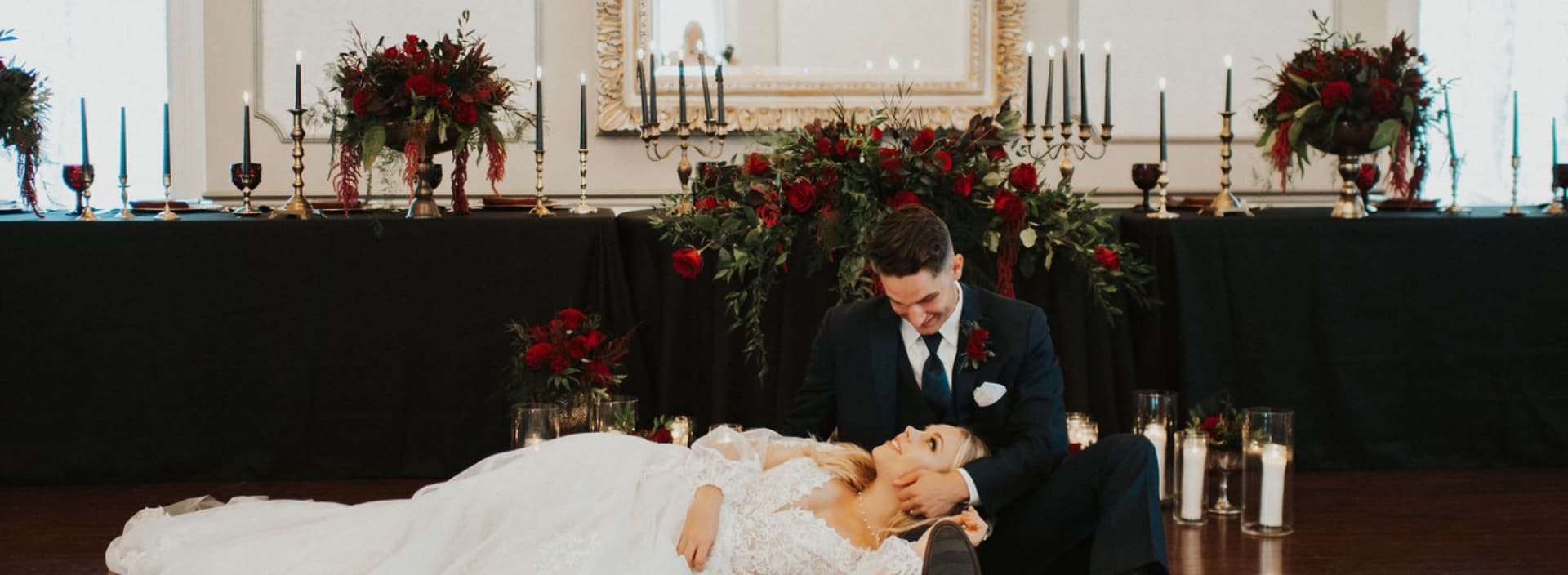 A groom sits next to his bride on the floor, surrounded by elegant floral arrangements and candles at a wedding reception.