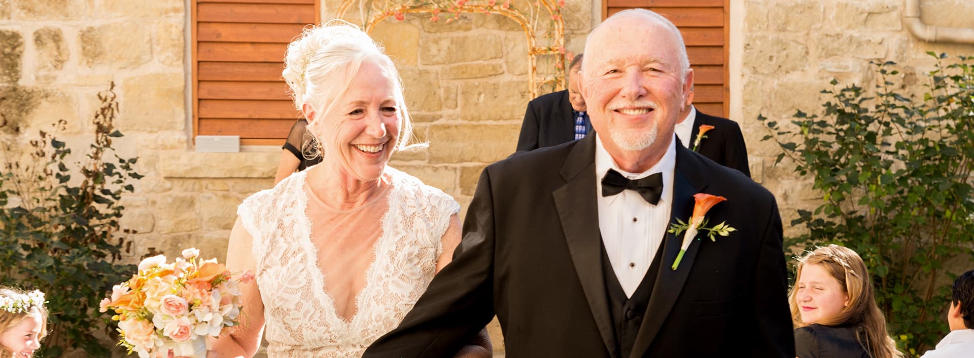 A smiling bride and groom walk together outdoors, surrounded by greenery and a rustic stone backdrop.