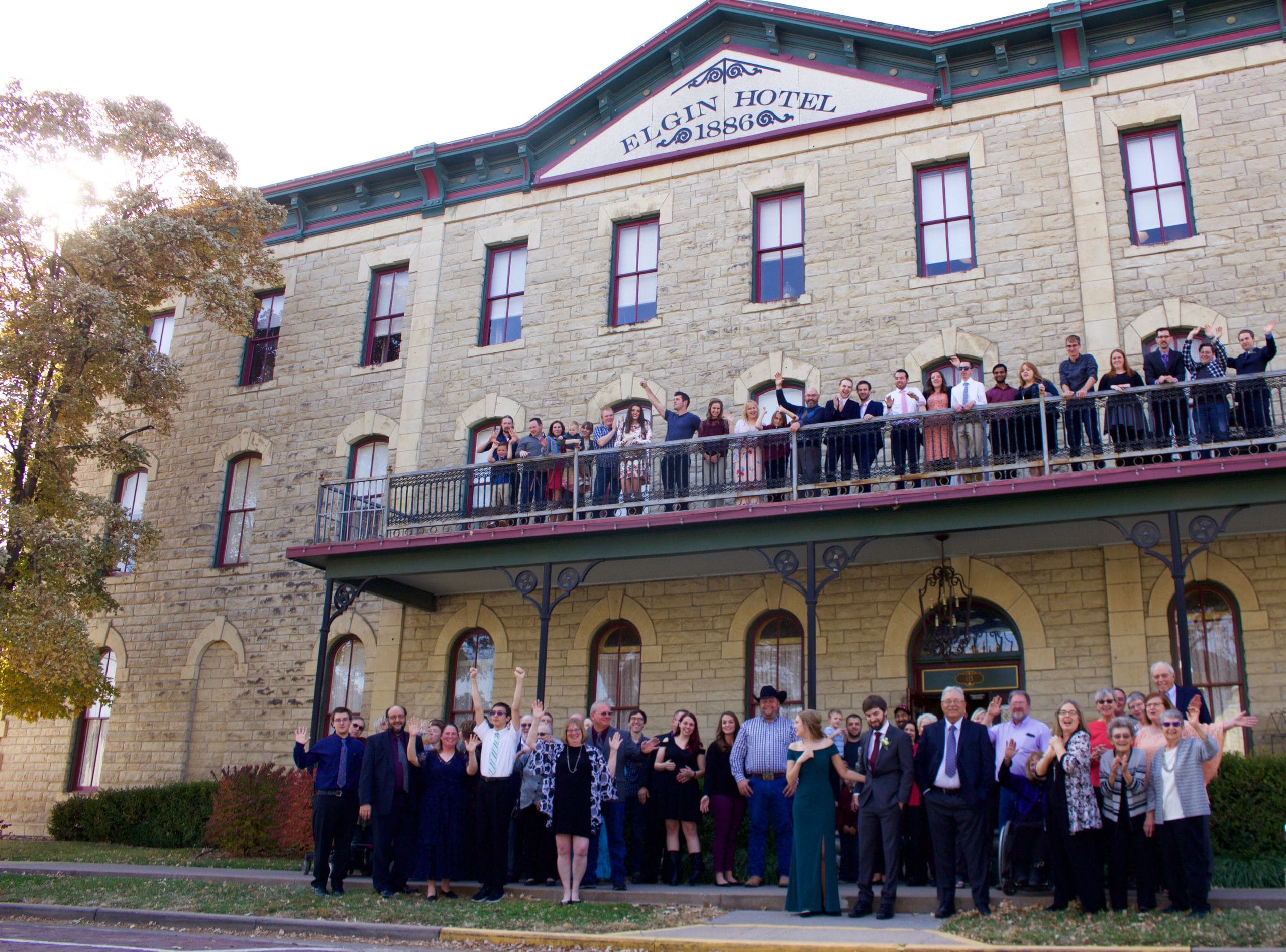 A large group of people joyfully wave from the balcony and ground level of the historic Elgin Hotel.