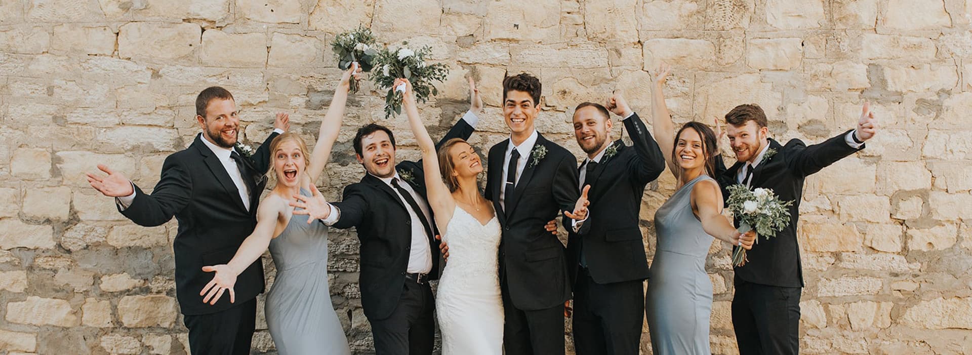A joyful wedding party poses enthusiastically in front of a stone wall.