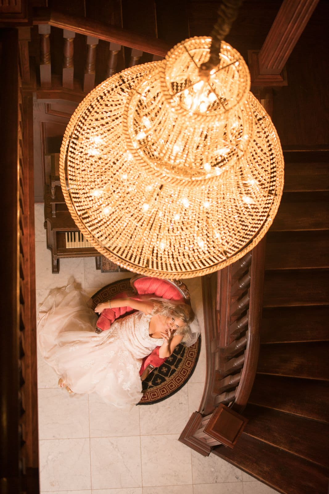 A bride rests elegantly on a plush chair beneath a grand chandelier in a staircase setting.