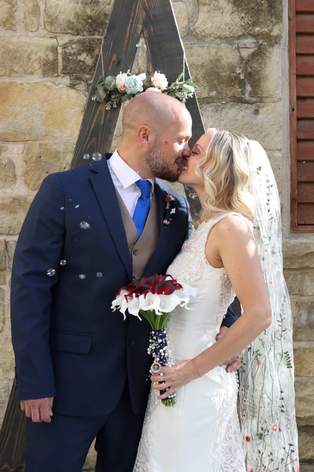 A bride and groom kiss surrounded by bubbles, with a floral arch and rustic stone background.