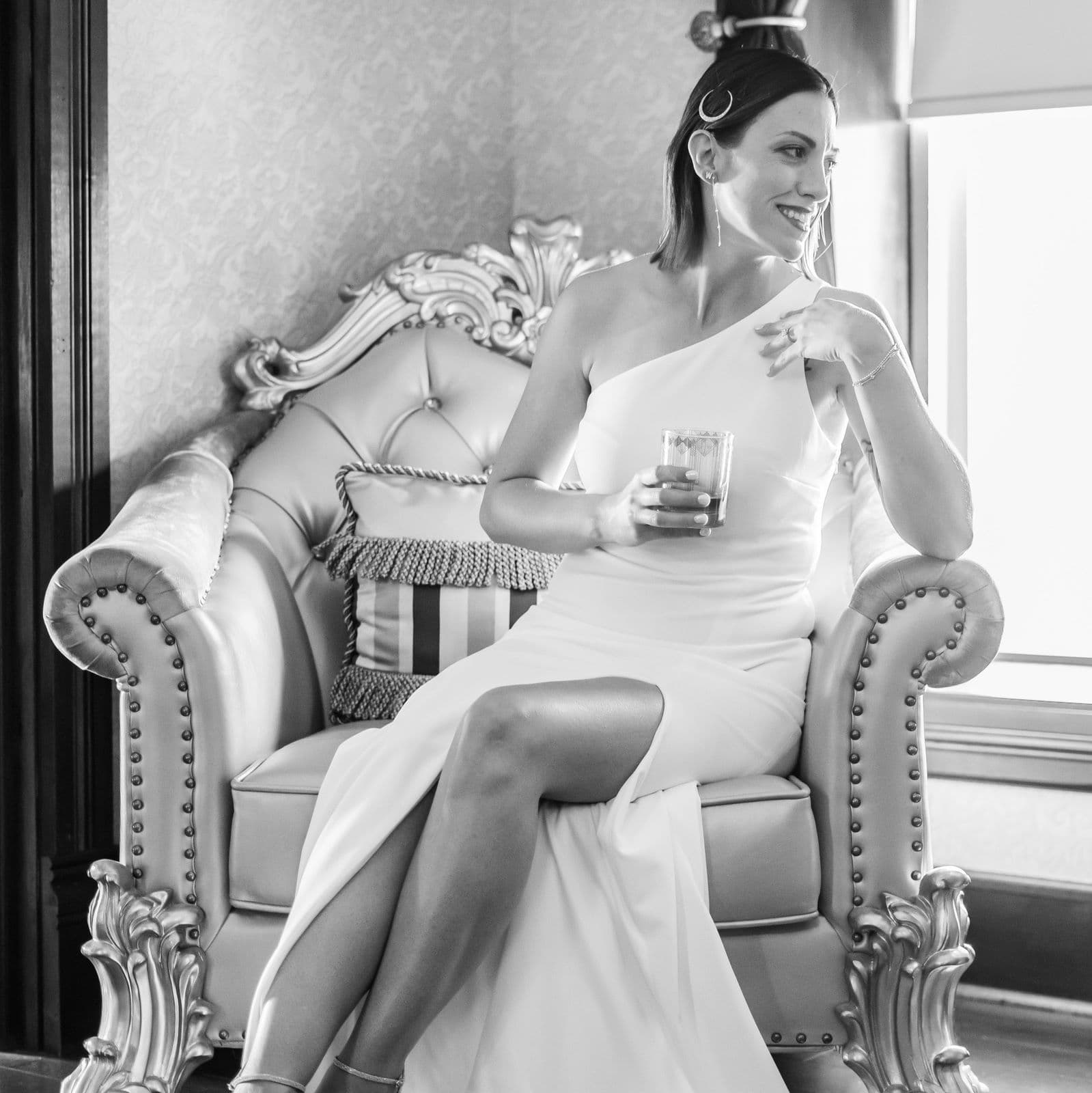 A woman in a white dress sits elegantly on a vintage chair, holding a drink and smiling.