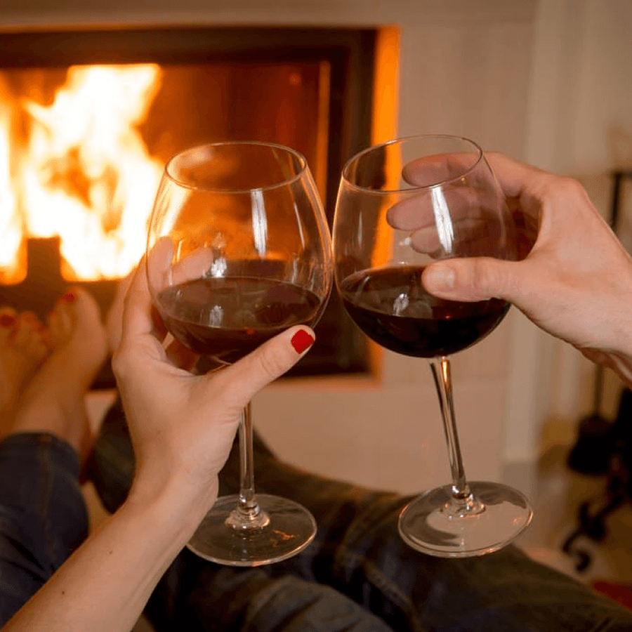 Two hands clink wine glasses in front of a cozy fireplace.
