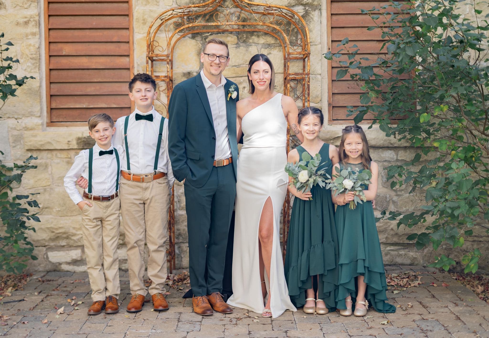 A couple in formal attire stands with four children, all dressed elegantly, against a rustic stone background.