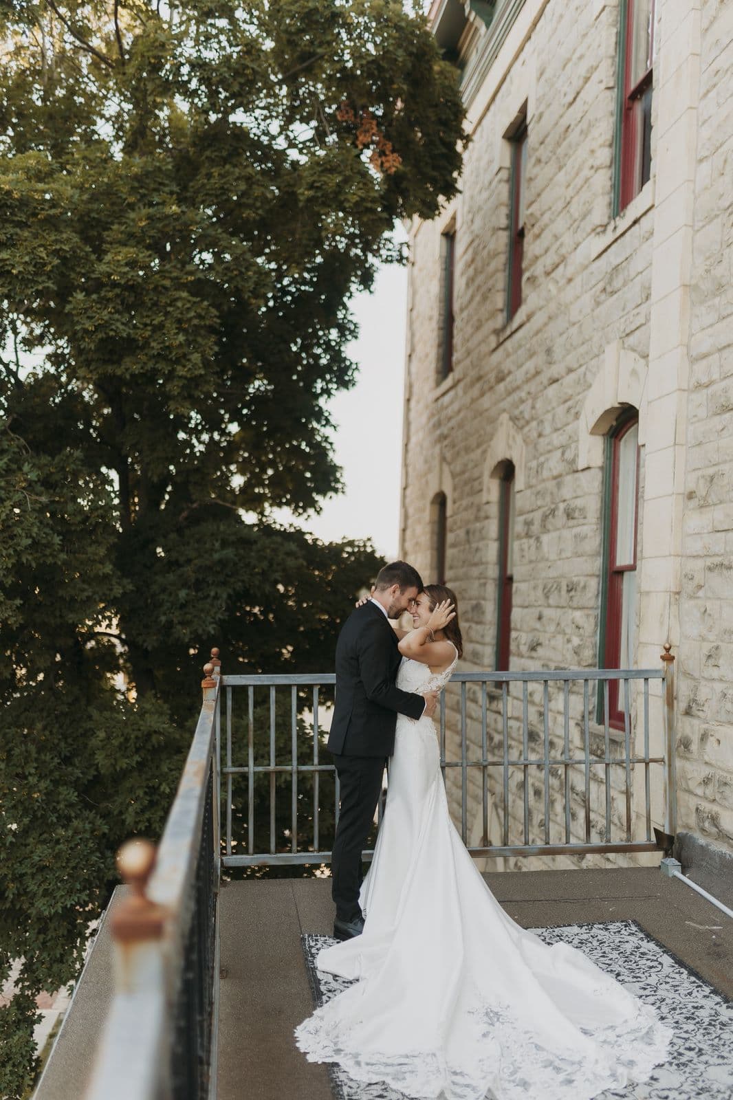 A couple embraces on a balcony surrounded by trees, dressed in wedding attire.
