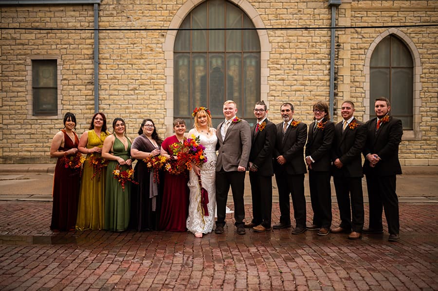 A bride and groom pose with their wedding party in front of a stone church on a rainy day.