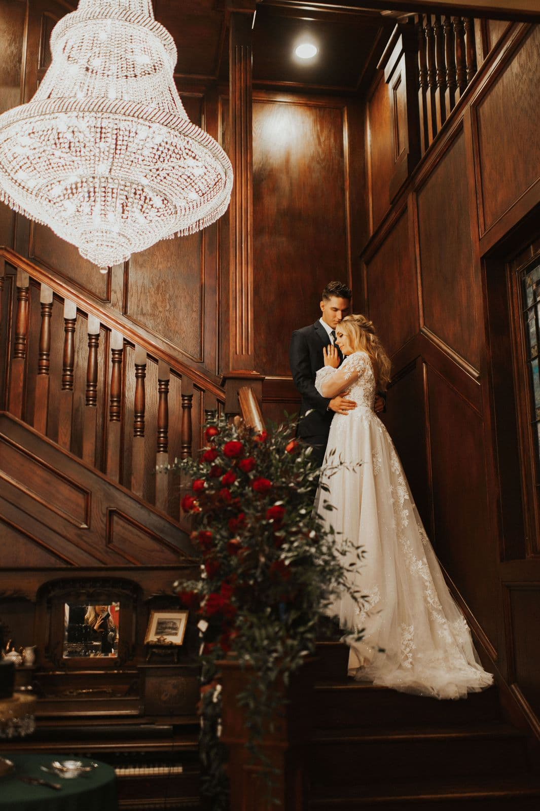 A newlywed couple embraces on a staircase adorned with a chandelier and floral arrangements.