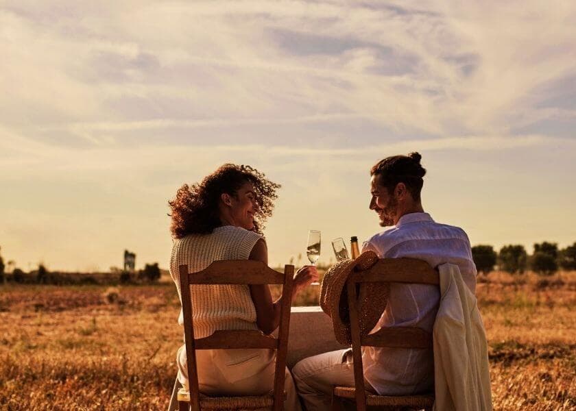 a smiling romantic couple facing away and toasting with wine while seated in wooden chairs at a table in the middle of an open field a smiling romantic couple facing away and toasting with wine while seated in wooden chairs at a table in the middle of an open field